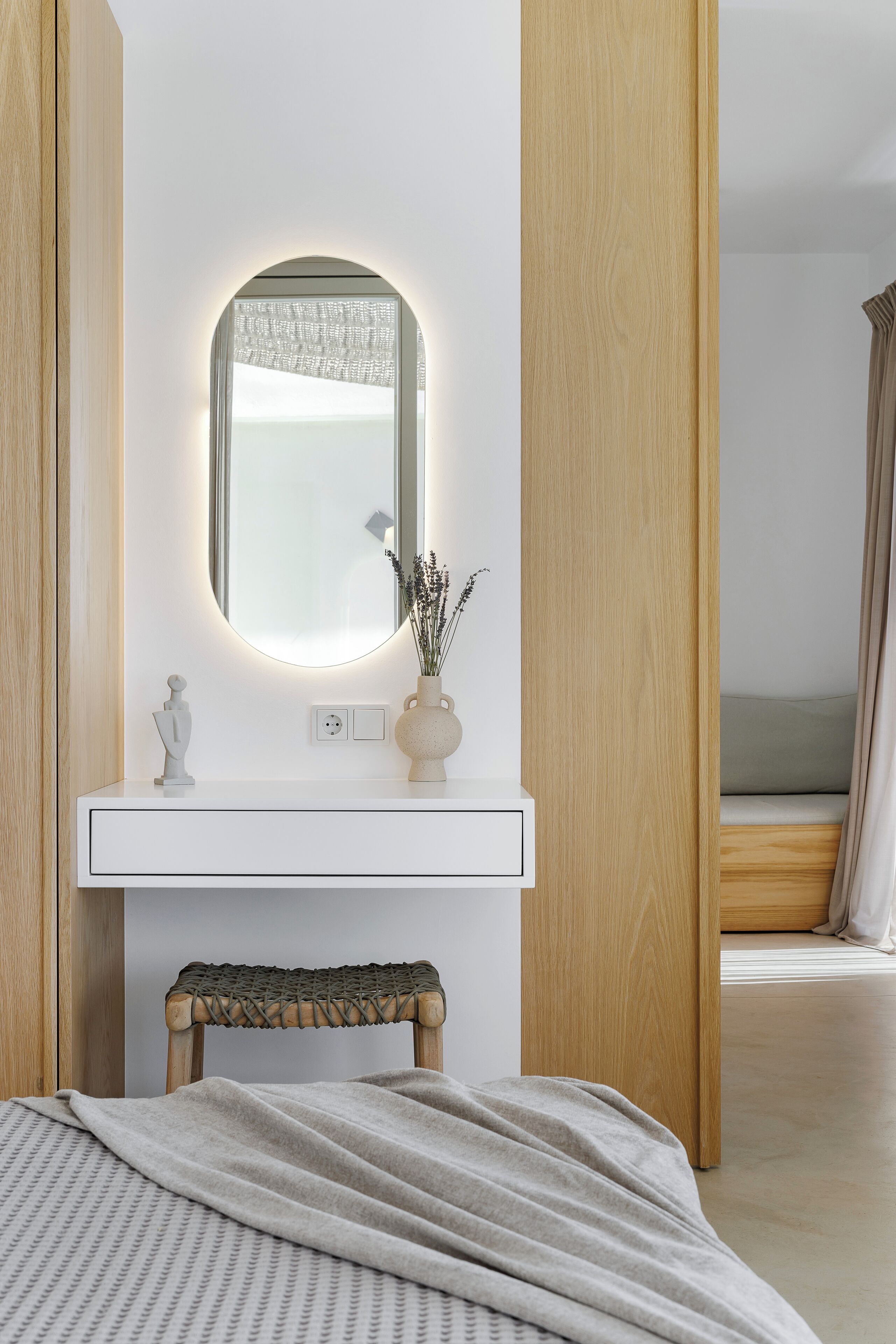 Un dormitorio moderno con un tocador de madera, un espejo redondo y una mesita de noche con un florero.