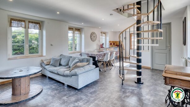 Ein geräumiges Wohnzimmer mit einem grauen Sofa, einem runden Holztisch und einer Wendeltreppe, die zu einer oberen Etage führt.