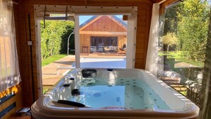 Eine Holzdiele mit Whirlpool in der Mitte, eine Holzterrasse davor und Blick auf ein Haus und Grünflächen.