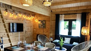 Ein Interieur einer Holzhütte mit einem für vier Personen gedeckten Esstisch, umgeben von Korbstühlen, einer Couch und einem an der Wand befestigten Fernseher.