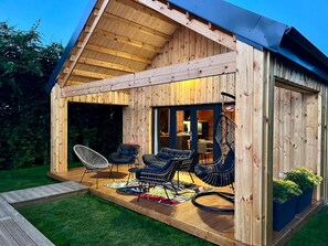 Une cabane en bois avec un patio couvert, deux chaises noires rembourrées, une chaise blanche et une chaise suspendue.