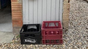 Een deur met een brievenbus en een meterkast, een zwarte kist met de tekst 'KONRAD', en een rode kist op een grindpad.