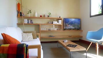 Un soggiorno moderno con un divano bianco, una coperta colorata, un tavolino in legno, una poltrona blu e un televisore su una mensola di legno.