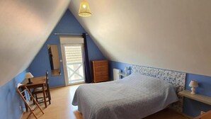 Een slaapkamer met een schuine kap, een bed met een grijze plaid, een houten dressoir, een nachtkastje, een lamp en een raam met uitzicht.