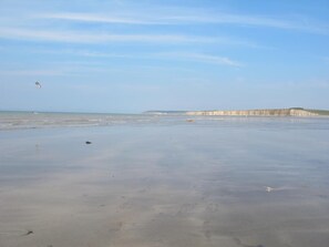 Une plage avec un ciel dégagé, une mer calme et des falaises qui se dessinent à l’horizon.
