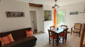 Een woonkamer met een donkerbruine bank, een houten salontafel en een eettafel met vier stoelen. Er is een keuken met een raam en een deur naar een andere kamer.