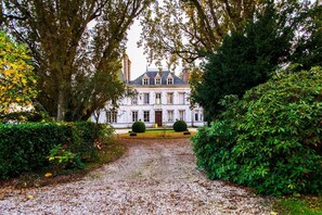 Un grand bâtiment symétrique, doté d’une entrée centrale et entouré de haies et d’arbres bien entretenus.