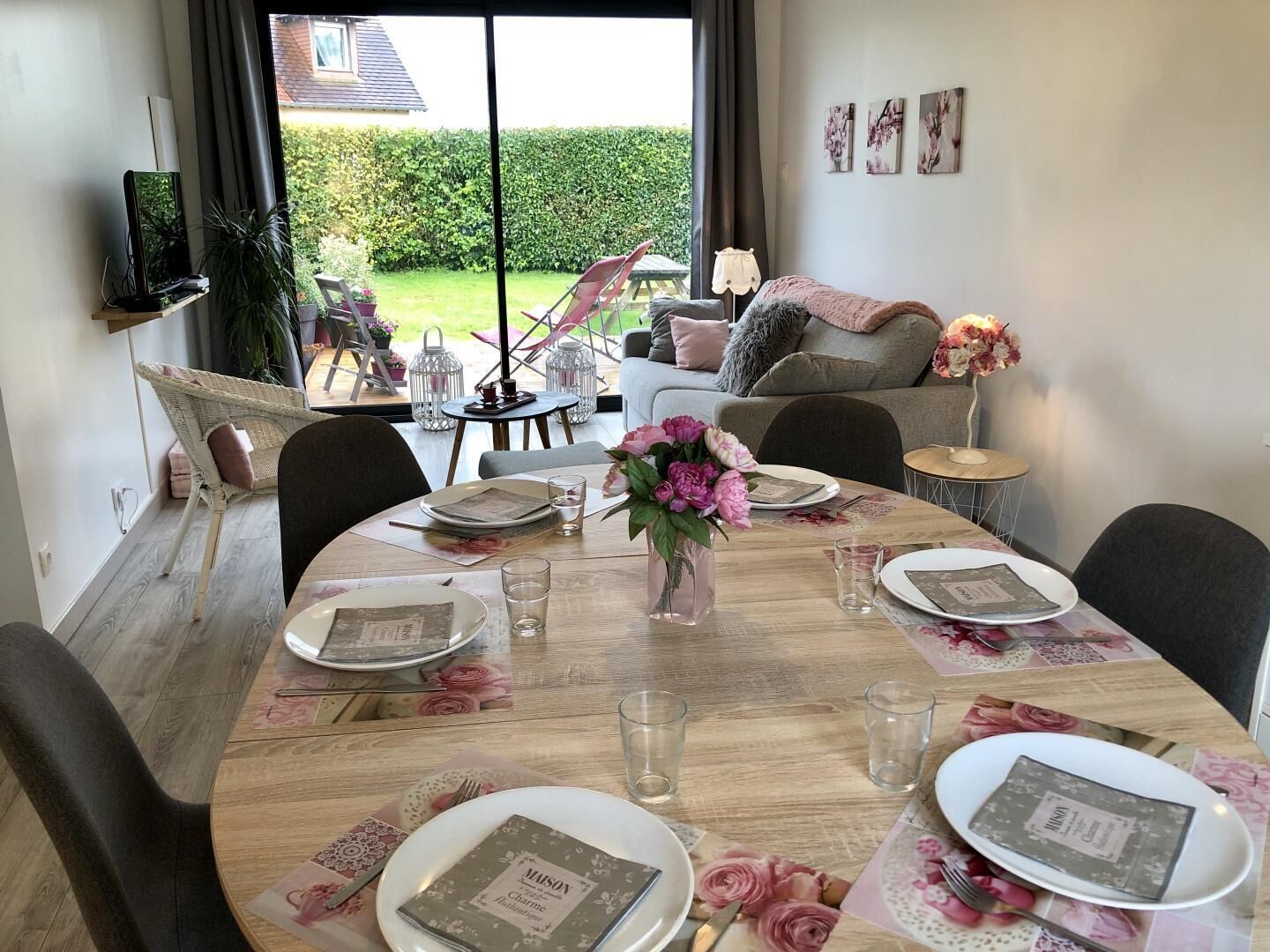 A dining area with a round table set for four, surrounded by chairs. There is a vase with flowers on the table, and the room has a view of a garden through large windows.