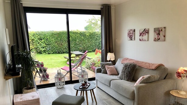 Un salon avec un canapé gris, une petite table, un banc et un téléviseur posé sur une étagère. Une grande fenêtre offre une vue sur un jardin.