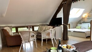 Une pièce confortable avec un lit, deux fauteuils en osier, une petite table avec une bouteille et des fruits, et un téléviseur.