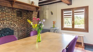 A dining area with a wooden table, purple chairs, a brick fireplace, and a window with a view of greenery. - Le Bremontier - Massy (MASSY)