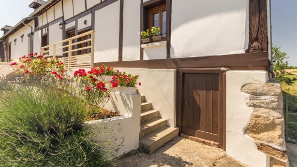 A traditional building with a white facade, dark wooden beams, and a stone base. There are steps leading to a wooden door, and potted plants with red flowers are visible. - Le Bremontier - Massy (MASSY)