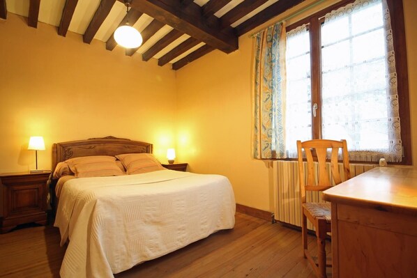 A bedroom with a bed, two bedside lamps, a wooden chair, a table, and a window with patterned curtains.