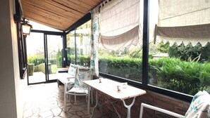 A covered patio with a stone floor, a wooden ceiling, and large windows overlooking a garden. - Le Rêve - Preaux - Gîte (PREAUX)