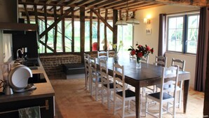 Un coin repas rustique avec une table en bois et des chaises blanches, un banc en pierre et une vue sur la verdure grâce à de grandes fenêtres.