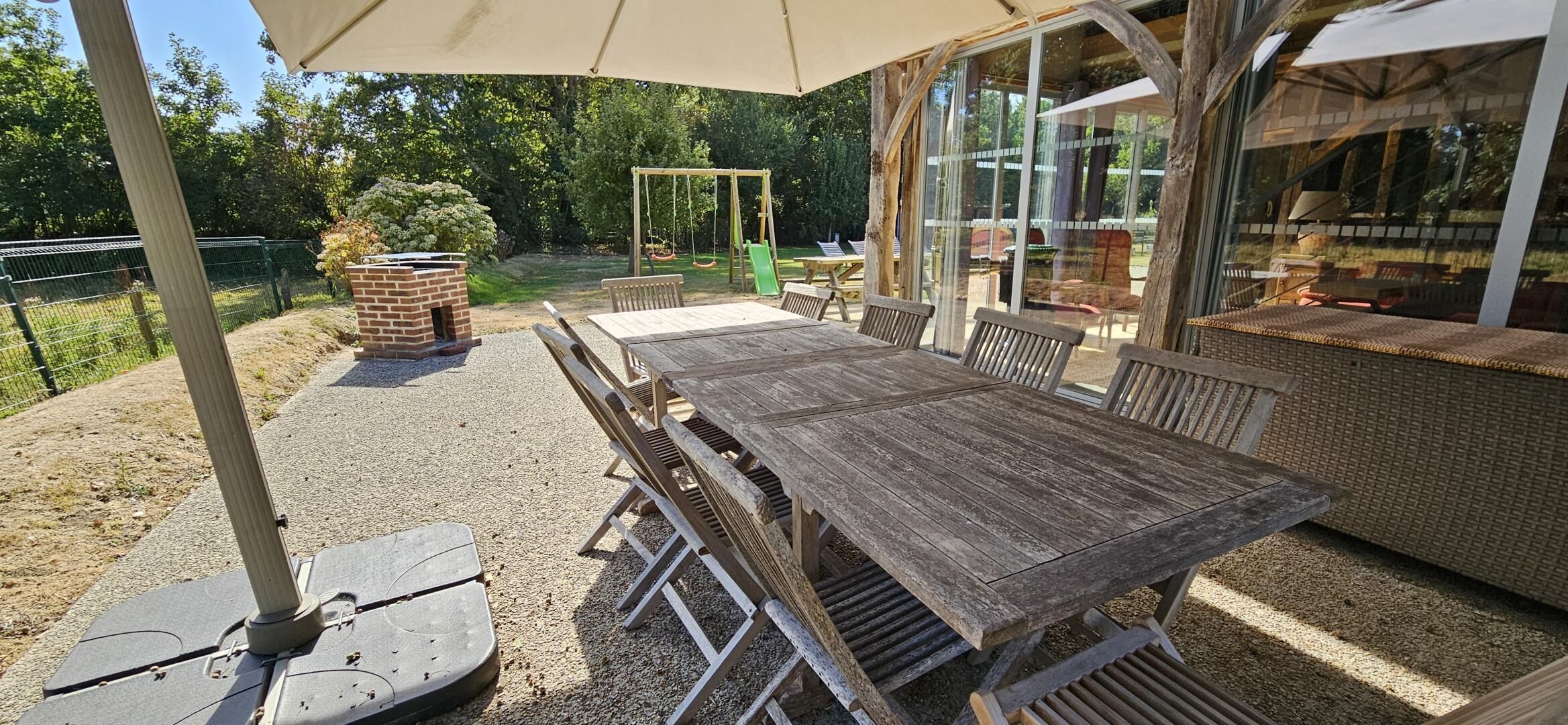 Een terras buiten met een houten tafel en stoelen, een bakoven en een schommel.