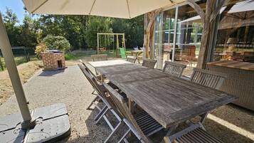 Een terras buiten met een houten tafel en stoelen, een bakoven en een schommel.