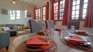 Ein Essbereich mit einem Holztisch, gedeckt für vier Personen, umgeben von Stühlen, mit einer Couch und einem Fenster mit roten Vorhängen im Hintergrund.