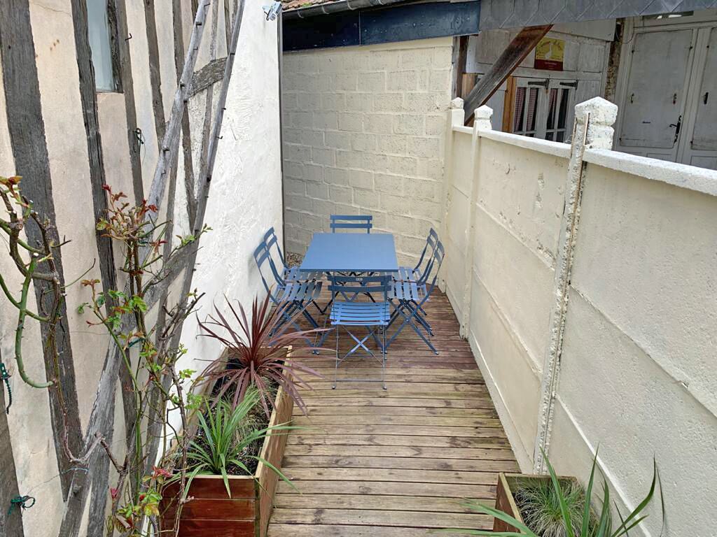 Un petit patio extérieur, avec une table et des chaises en bois, entouré de plantes en pot et bordé d’un mur blanc.