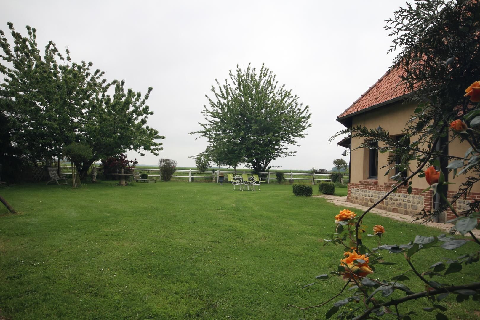 A well-maintained grassy yard with a white fence, a building with a red roof, and a few trees.