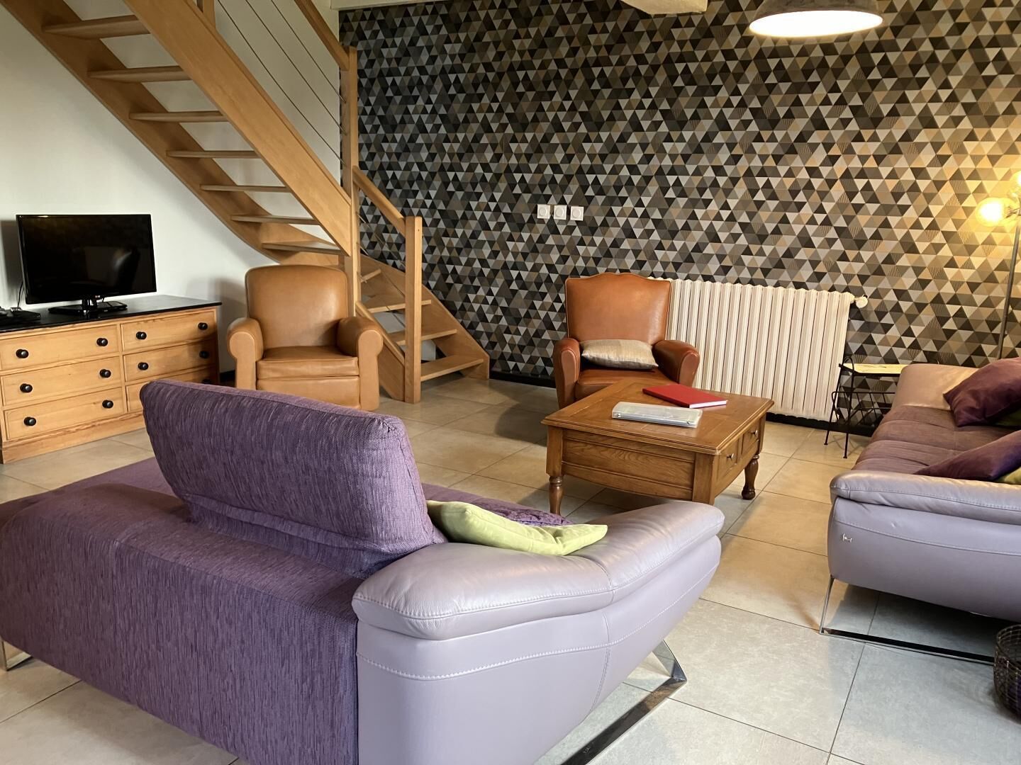 Un salon moderne avec un canapé violet, une table basse en bois, un fauteuil en cuir et un escalier agrémenté d’un mur aux motifs géométriques.