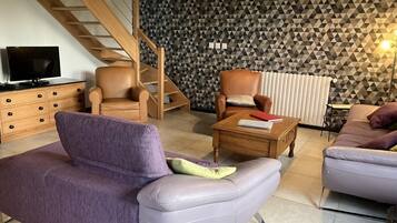 Un salon moderne avec un canapé violet, une table basse en bois, un fauteuil en cuir et un escalier agrémenté d’un mur aux motifs géométriques.
