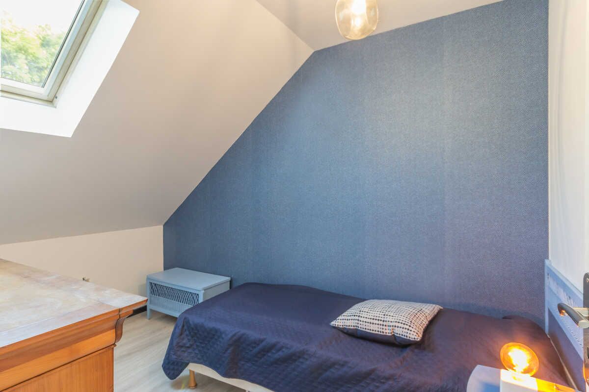 A bedroom with a sloped ceiling, a bed with a dark blue bedspread, a bedside table, a small nightstand, and a lamp.