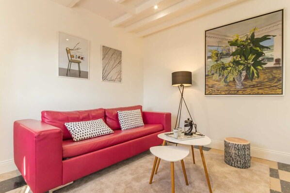 Una sala de estar moderna con un sofá de cuero rojo, una mesita blanca, una lámpara de pie y un cuadro de plantas.