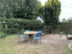 Una mesa redonda de madera con cuatro sillas azules en un jardín con abundante follaje y un árbol alto.