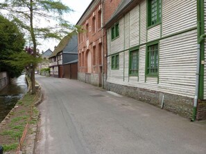 A narrow street lined with historic buildings, including a brick structure with arched windows and a white building with green window frames. - Gîtes de France® - Le Héron (ST SAENS)