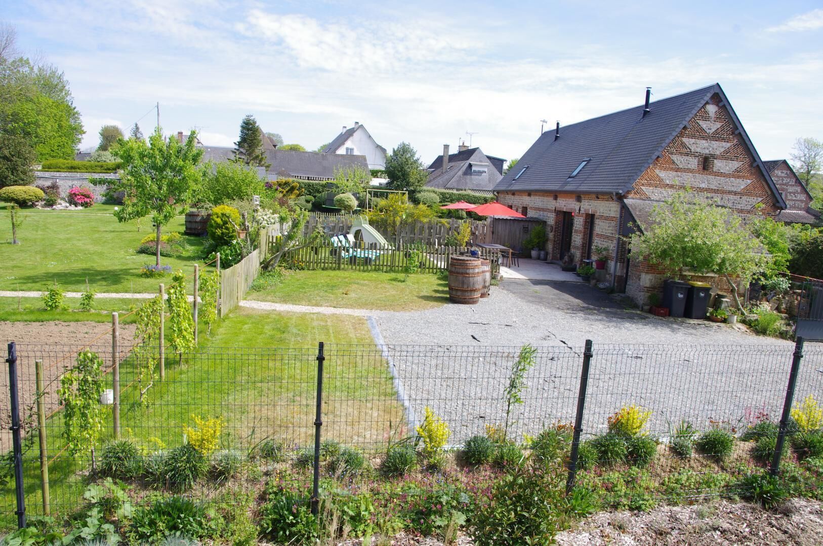 A rural property with a fenced garden, a wooden play structure, and a brick building with a sloped roof.