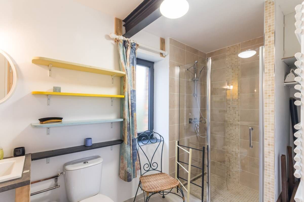 A bathroom with a toilet, sink, shower, and a chair. Shelves are mounted on the wall, and a window is visible.