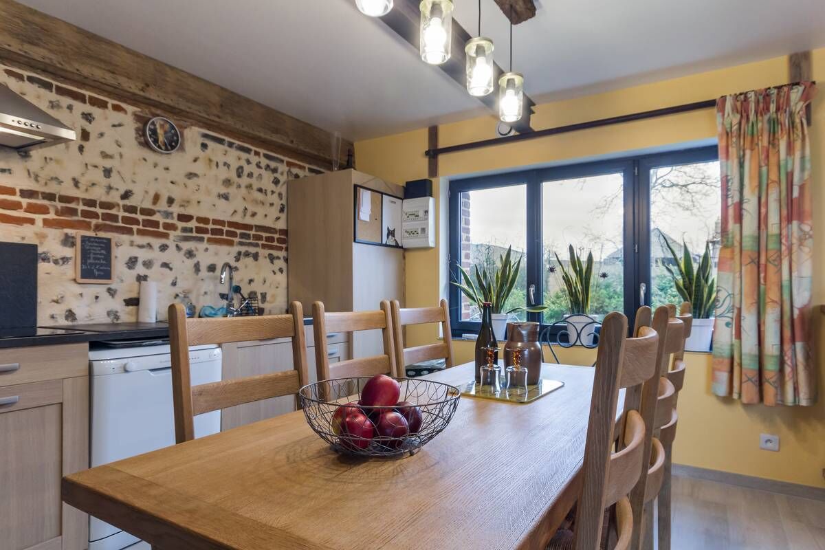 Una cocina con pared de ladrillo, sillas de madera, una mesa de comedor con frutero y una ventana con plantas.