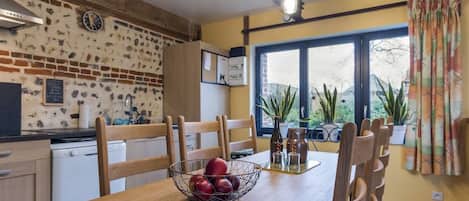 Una cocina con pared de ladrillo, sillas de madera, una mesa de comedor con frutero y una ventana con plantas.
