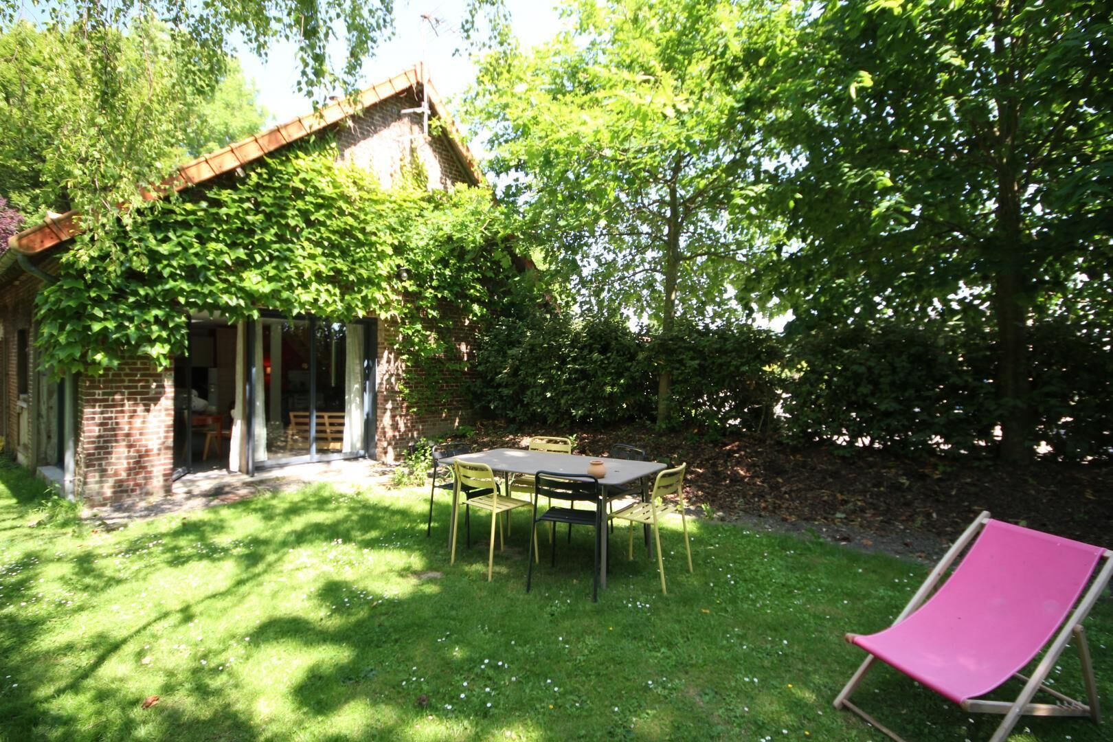 Een terras met een tafel en stoelen, omringd door weelderige begroeiing en een bakstenen gebouw.