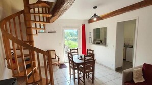 A spacious living area with a wooden staircase, a dining table with chairs, a sofa, and a kitchen area visible through an open door. - Rose - Bourg DUN (BOURG DUN (LE))