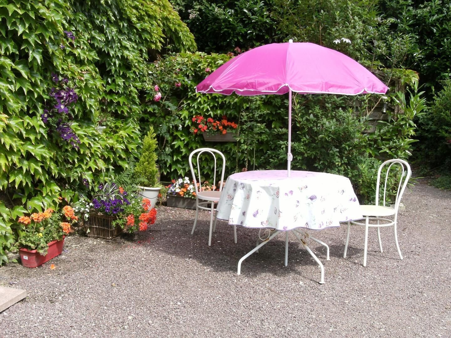 Ein Garten mit Tisch und Stühlen unter einem rosa Sonnenschirm, umgeben von verschiedenen Blumentöpfen und Blumen.