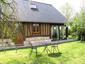 Una casa con base de piedra, vigas de madera y techo inclinado. Cuenta con un patio con mesa y sillas, y un espacio cubierto con puerta de vidrio.