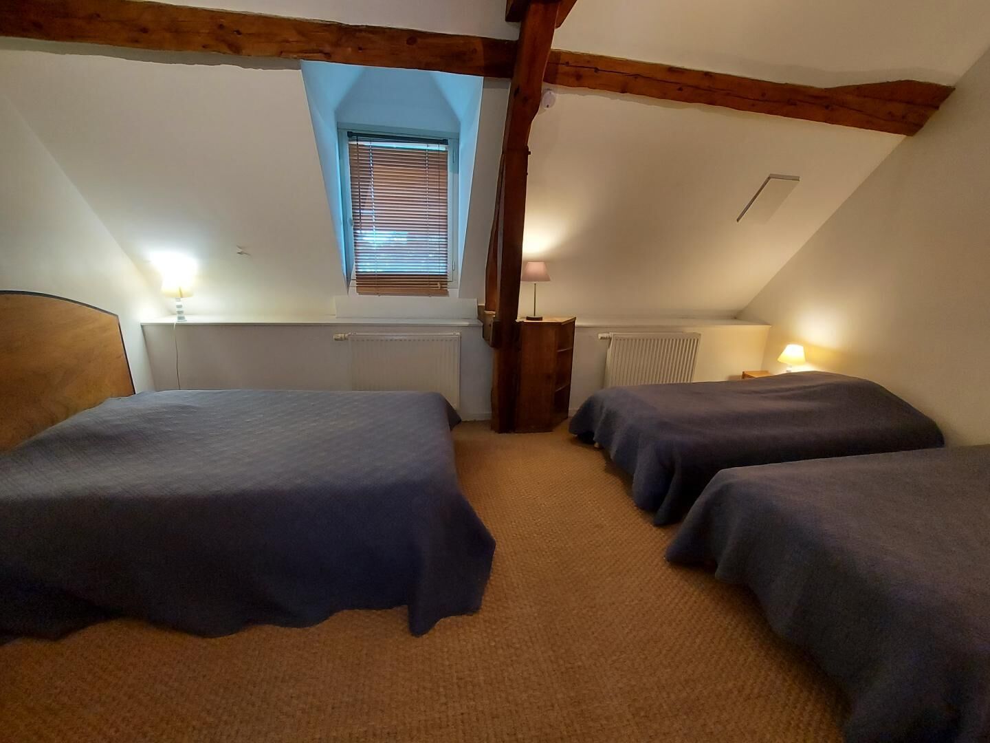 A room with two beds, a window with blinds, wooden beams, and a radiator.