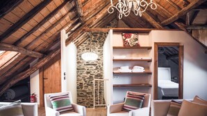 Ein gemütliches Dachzimmer mit Holzbalken, einer Steinwand und einem Bett. Zwei weiße Sessel mit gestreifter Polsterung, ein kleiner Tisch und eine Pendelleuchte sind ebenfalls vorhanden.