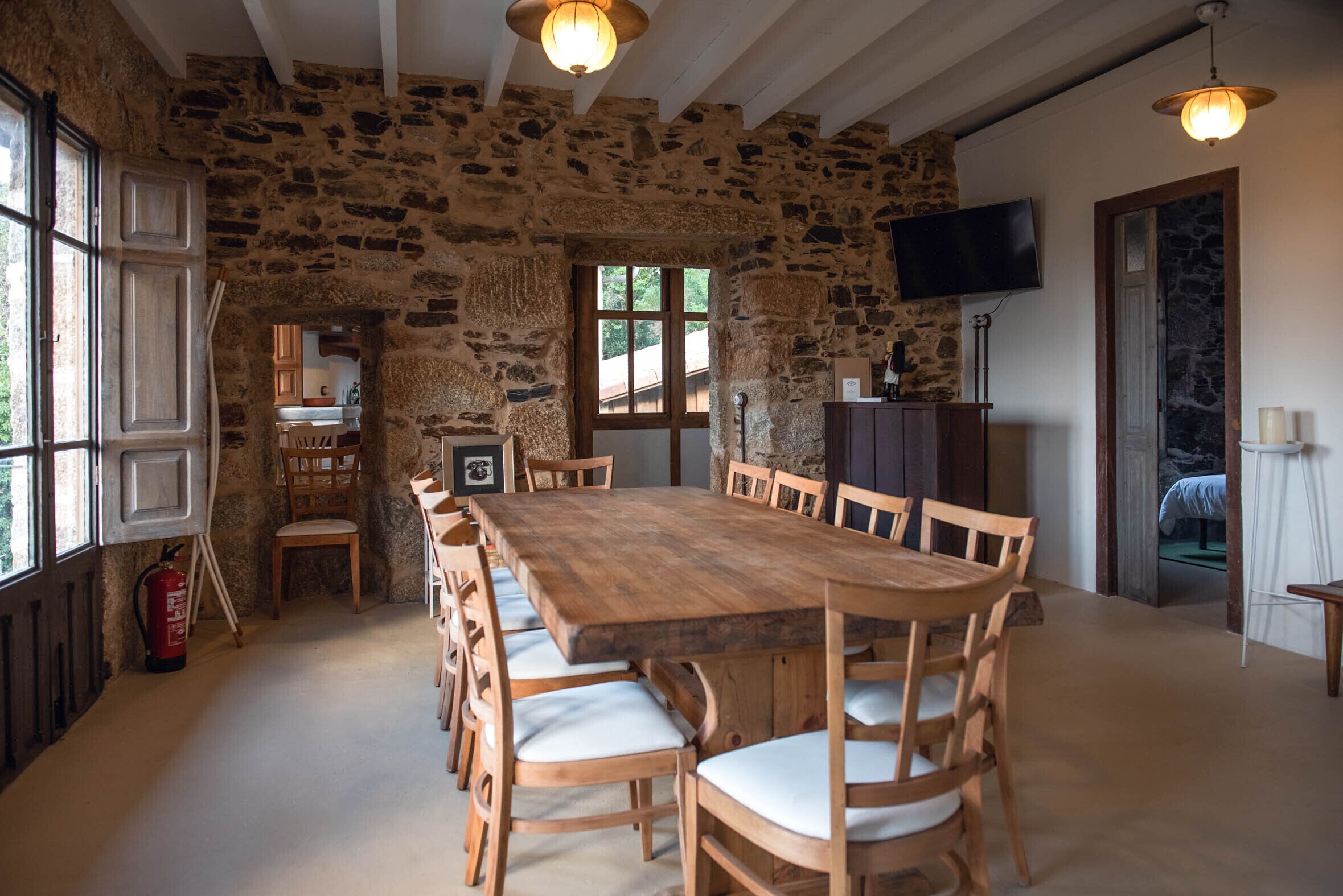 Ein Essbereich mit Steinwänden, einem langen Holztisch und Stühlen, einem an der Wand befestigten Fernseher und einem Feuerlöscher in der Nähe der Fenster.