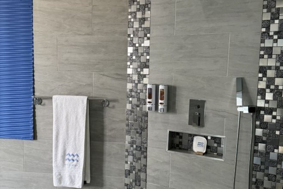 A modern bathroom with grey tiles, a blue towel, a soap dispenser, and a showerhead.