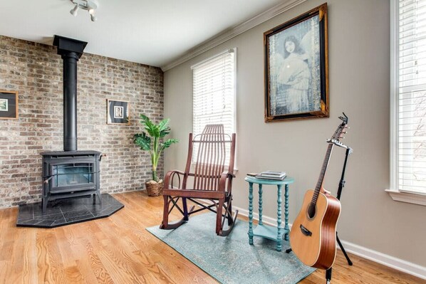 Un salon chaleureux avec un mur en briques, une cheminée, un fauteuil à bascule, une guitare et une fenêtre avec des stores.