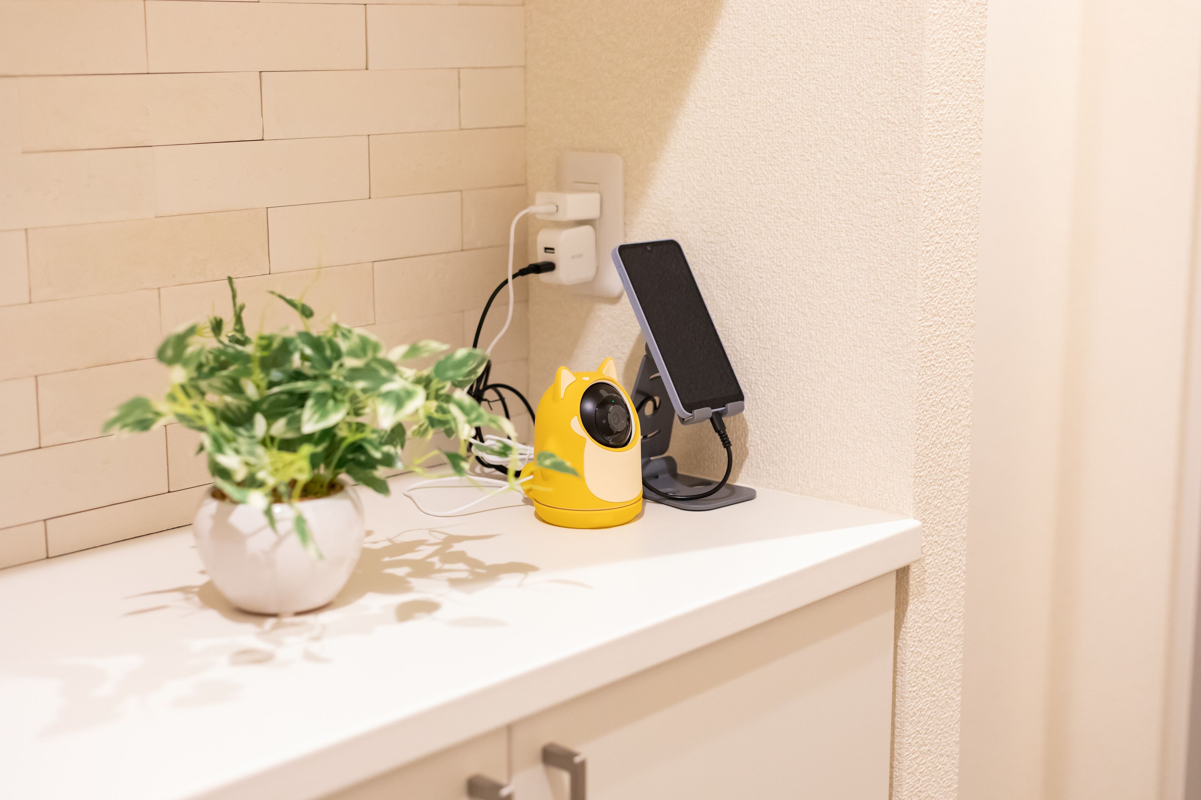 Una encimera blanca con una planta en maceta, un dispositivo amarillo y un teléfono inteligente sobre un soporte.