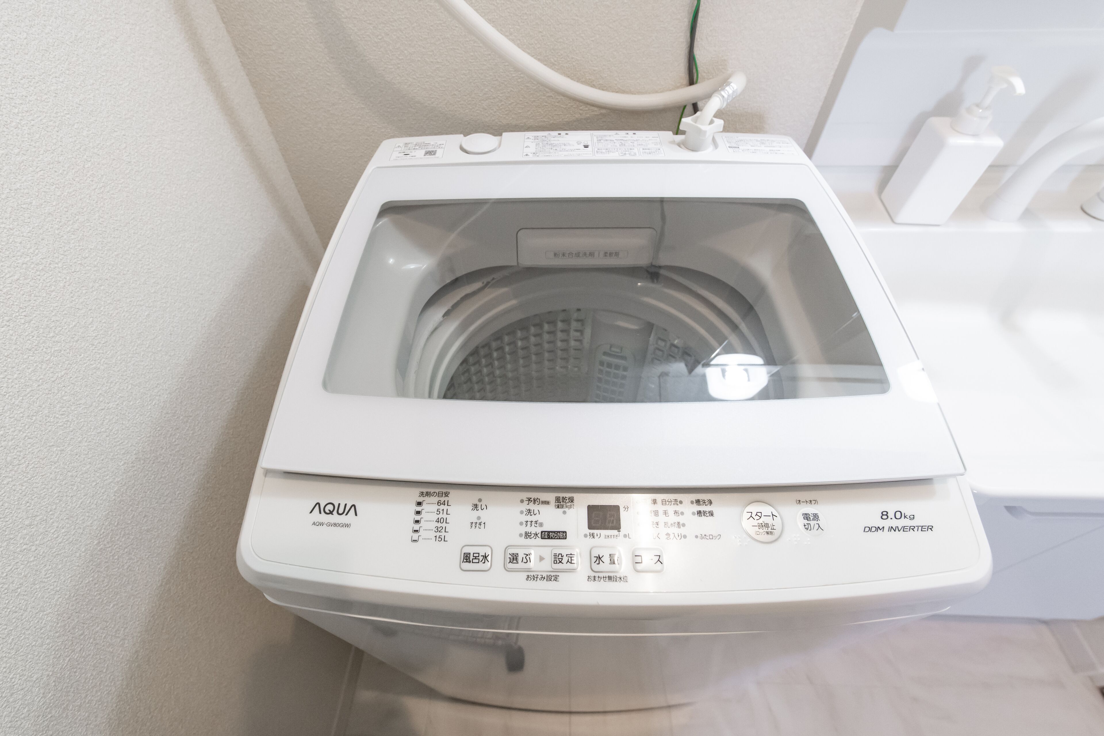 A front view of a white front-loading washing machine with a control panel, a water inlet hose, and a white wall in the background.