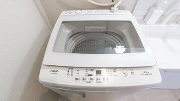 A front view of a white front-loading washing machine with a control panel, a water inlet hose, and a white wall in the background.