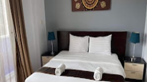 A neatly made bed with white linens and two rolled towels, a wooden headboard, and two bedside lamps on nightstands. - LaXenia (Karon)