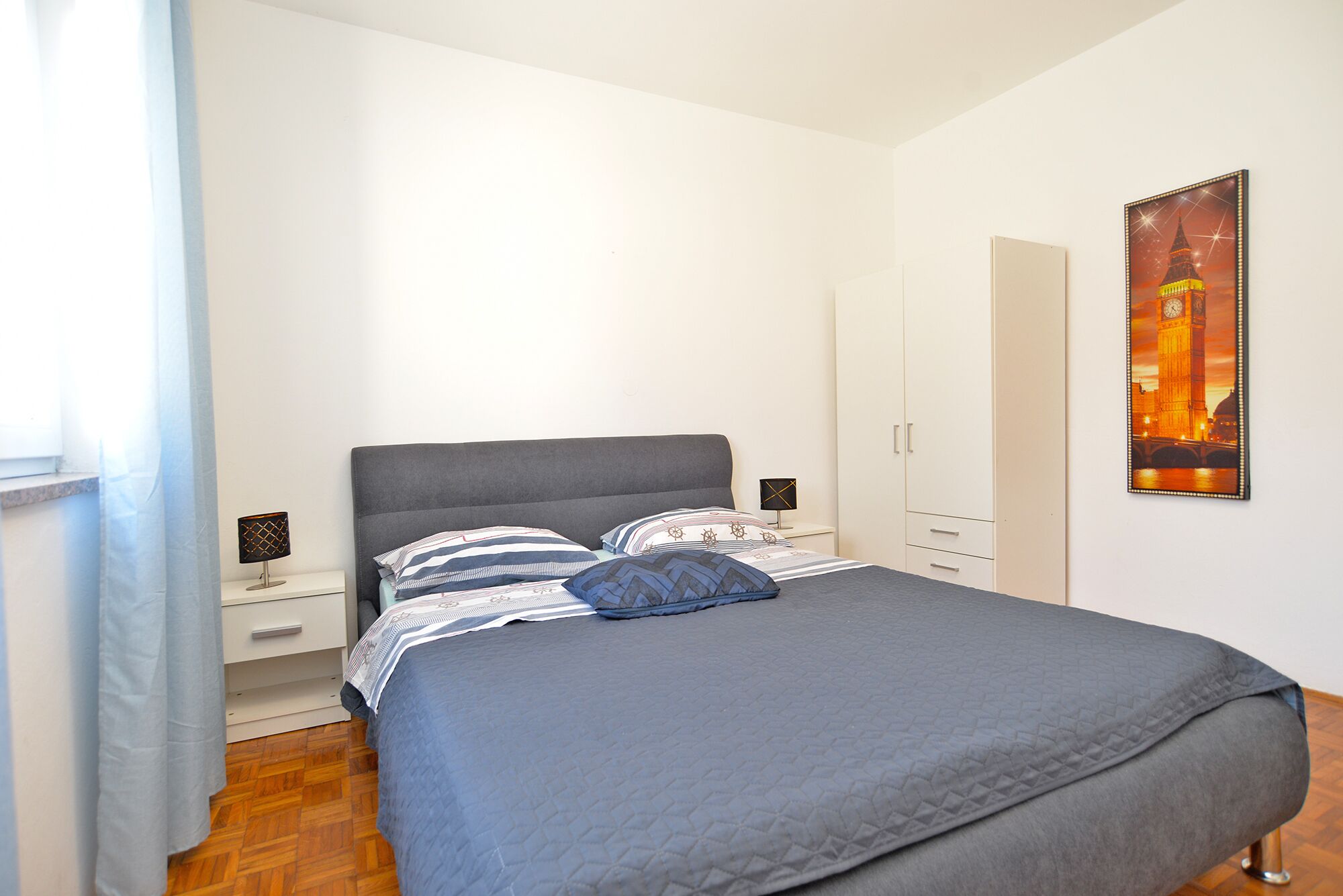A bedroom with a bed, nightstands, a wardrobe, and a framed picture of a famous landmark.
