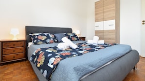 A neatly made bed with a floral bedspread, a grey quilted duvet, and white towels. There is a wooden nightstand with a lamp, a dark grey upholstered headboard, and a wooden wardrobe with two drawers.