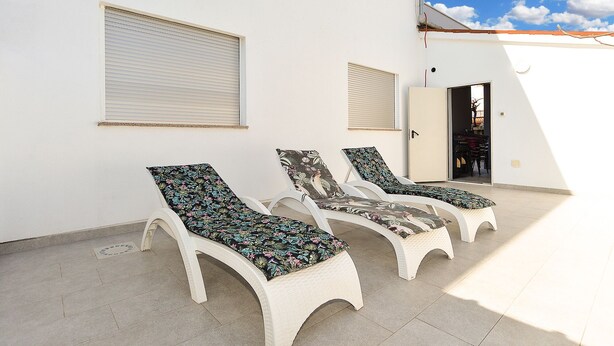 A white building with three white lounge chairs with floral cushions, a door, and a window with blinds.